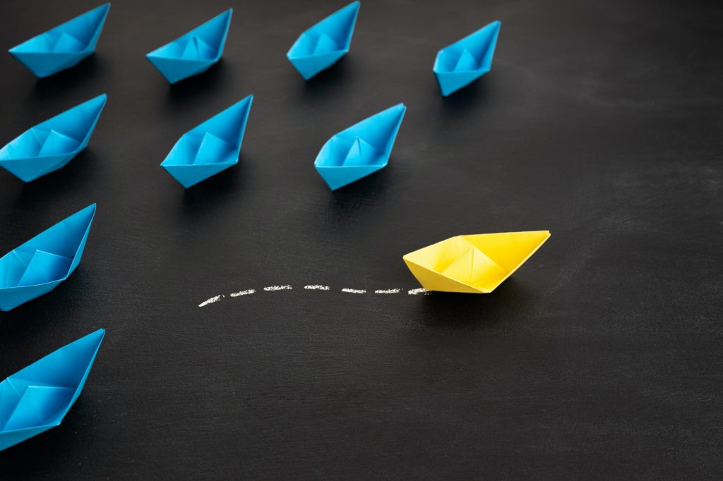 A group of blue paper boats with a single yellow boat forging its own path.